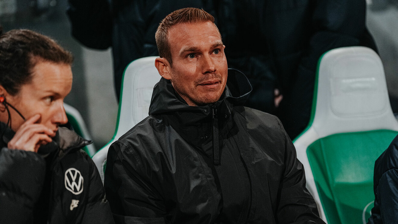 VfL-Wolfsburg-Trainer Tommy Stroot sitzt auf der Bank und lächelt.