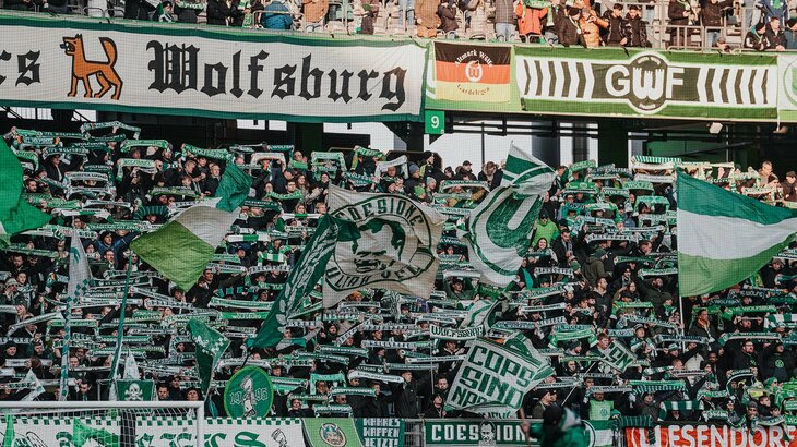 Fans des VfL Wolfsburg stehen auf der Tribüne und halten grün-weiße Schals hoch.