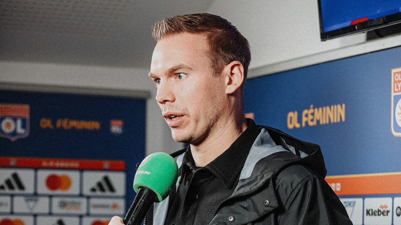 VfL-Wolfsburg-Frauen-Trainer Tommy Stroot steht vor einer Werbetafel und hat ein Mikrofon vor seinem Mund.