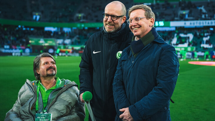 Ein ALS-Erkrankter sitzt neben dem Stadionsprecher vom VfL Wolfsburg und Roy Präger im Rollstuhl. Sie stehen an der Seitenlinie in der Volkswagen Arena.