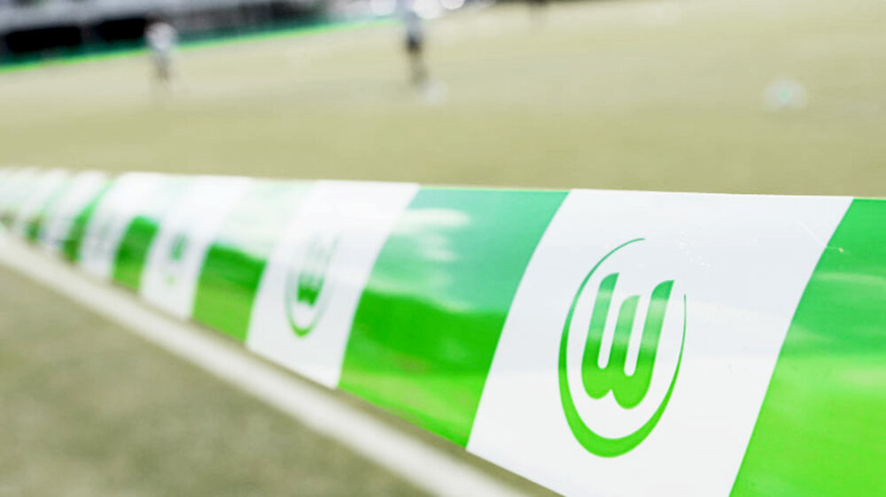 Ein VfL Wolfsburg-Flatterband ist um Vordergrund, im Hintergrund erkannt man unscharf einen Fußballplatz. 