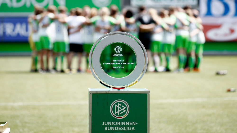 Im Vordergrund ist die Meisterschale, im Hintergrund bilden die Spielerinnen des VfL Wolfsburg einen Kreis.