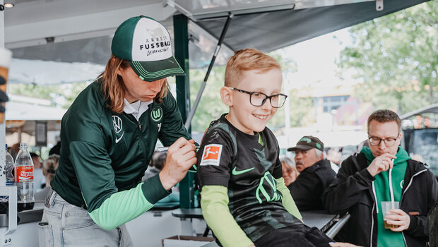 Patrick Wimmer vom VfL Wolfsburg unterzeichnet auf dem VfL Trikot einen kleinen Jungen. 