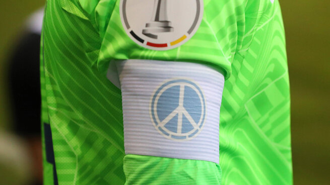 Nahaufnahme einer Kapitänsbinde mit Peace Zeichen am Arm eines VfL Wolfsburg Spielers.