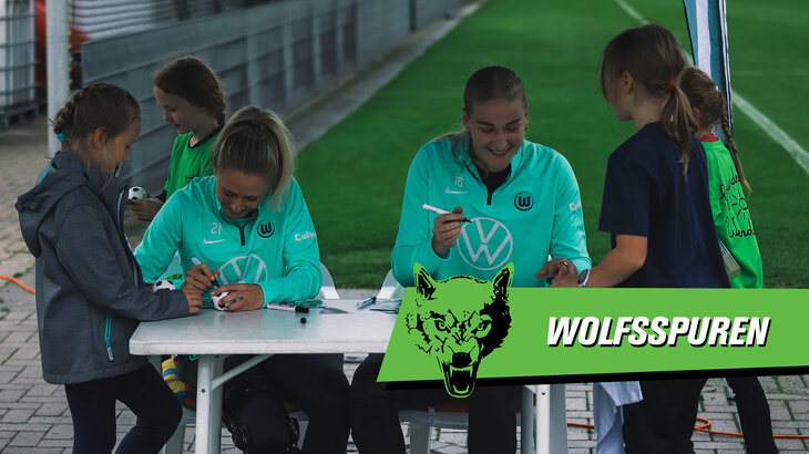 Eine Grafik mit dem Wolfsspuren-Logo. Im Hintergrund geben die VfL-Wolfsburg-Spielerinnen Rebecka Blomqvist und Camilla Küvers Autogramme.
