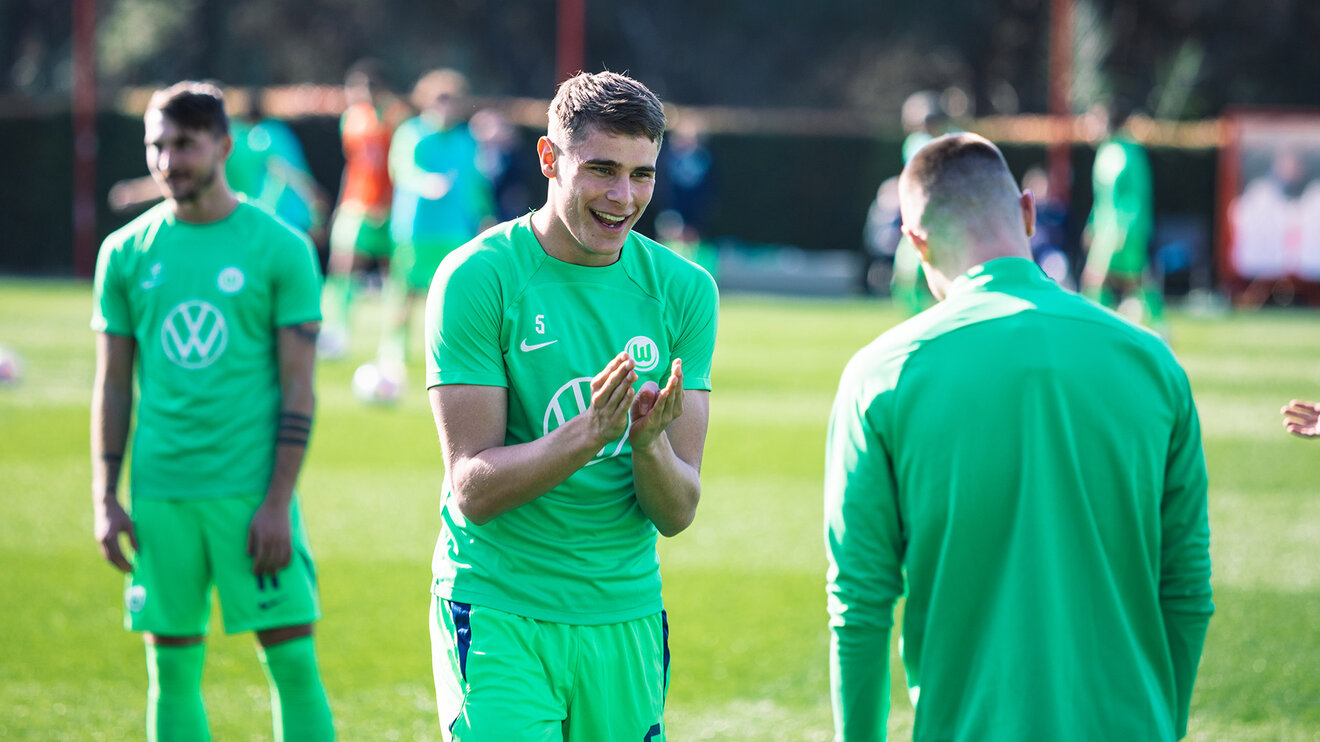 VfL-Wolfsburg-Spieler Micky van de Ven hält die Hände vor der Brust und lacht.