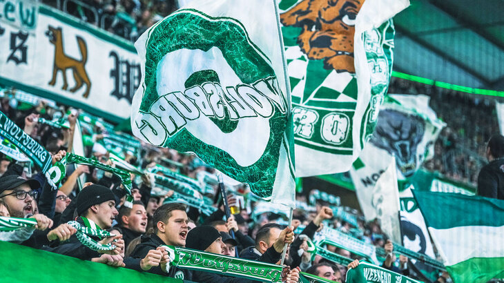 Die Fans des VfL Wolfsburg wedeln in der Fankurve mit ihren Fahnen und halten ihre Fanschals hoch.