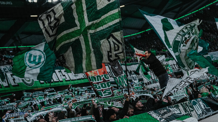 Die Fans des VfL Wolfsburg mit Fahnen und Schals mit Stadion auf der Tribüne.