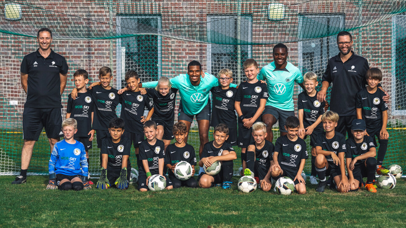 VfL Wolfsburg Spieler Jenz und Baku posieren mit der U10 eines VfL Partnervereins auf deren Trainingsplatz.