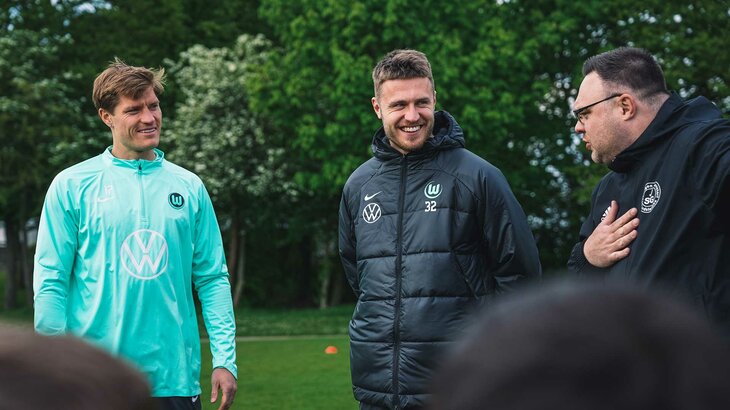 VfL-Wolfsburg-Spieler Mattias Svanberg und Kevin Behrens lachen während sie mit dem Trainer des Nordsteimker Nachwuchs sprechen.