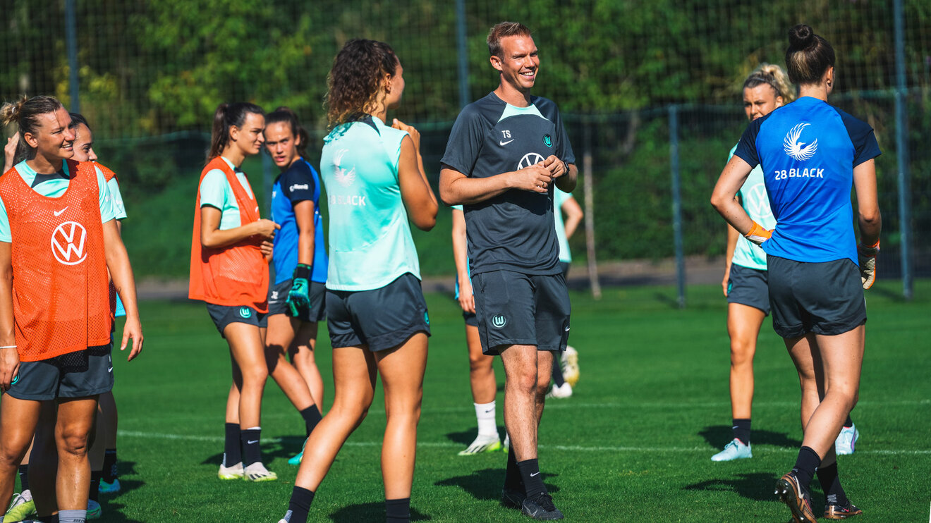 VfL-Wolfsburg-Trainer Tommy Stroot lachend mit Spielerinnen während einer Trainingseinheit.