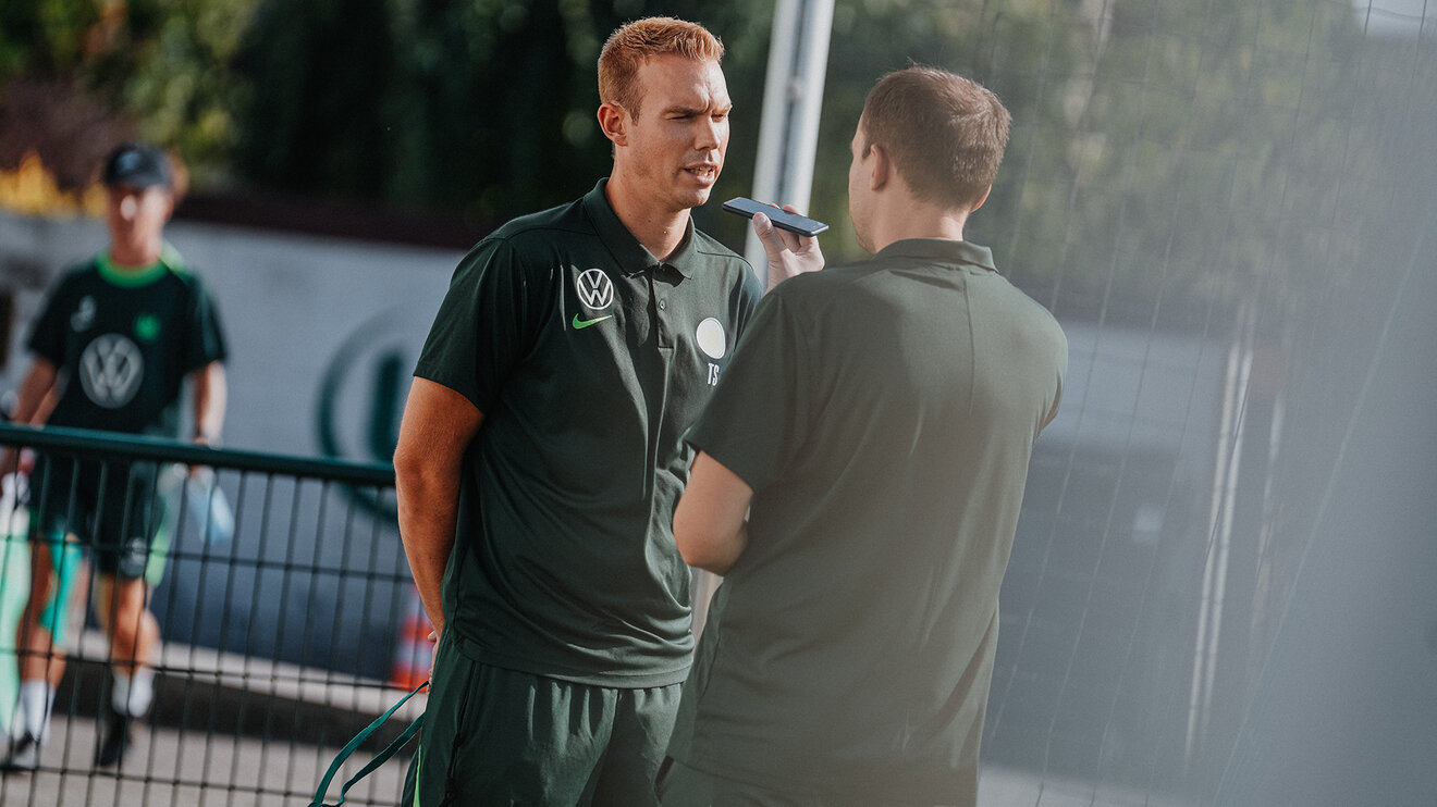 VfL-Wolfsburg-Frauentrainer Tommy Stroot steht in der Mitte. Vor ihm steht ein Mann der ihm ein Mikrofon entgegenstreckt.