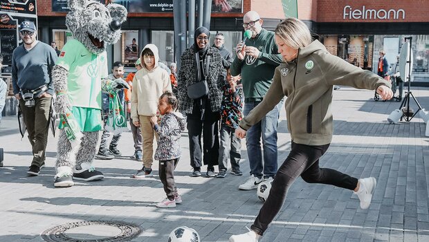 Kristin Demann vom VfL Wolfsburg kickt mit ihrem rechten Fuß den Ball in Richtung Torwand. Um Sie herum stehen mehrere Besucher und Wölfi und schauen zu.