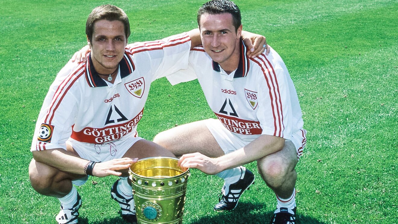 Marco Grimm hockt mit einem Mitspieler auf dem Rasen und fasst den DFB-Pokal an.
