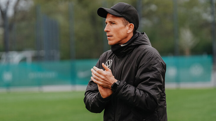 VfL-Wolfsburg-U19-Trainer Daniel Bauer steht am Spielfeldrand und klatscht.