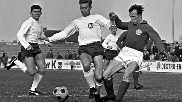 Ein schwarz-weiß Foto aus der Historie des VfL Wolfsburg zeigt zwei Spieler im Zweikampf um den Ball. Ein dritter Spieler beobachtet die Szene aus dem Hintergrund.