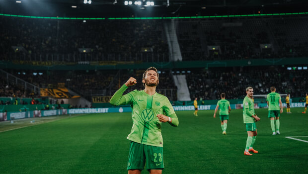 Ein VfL-Wolfsburg-Spieler streckt seinen linken Arm jubelnd in die Luft. Im Hintergrund sind weitere Spieler zu sehen und die Fankurve vom BVB.