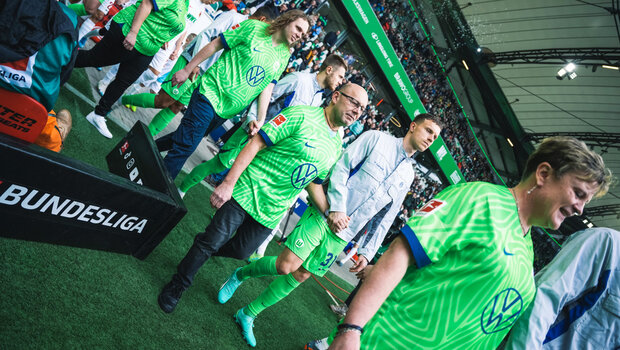 Die VfL-Wolfsburg-Spieler laufen in den Volkswagen-Arbeitsjacken mit Mitarbeitern auf das Spielfeld.