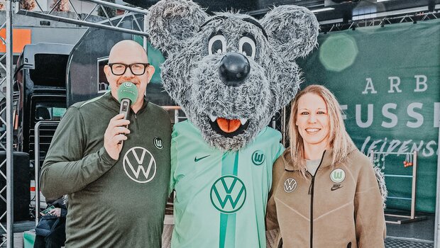 VfL Wolfsburg Moderator, Wölfi und Kristin Demann vom VfL Wolfsburg stehen nebeneinander und lächeln in die Kamera.