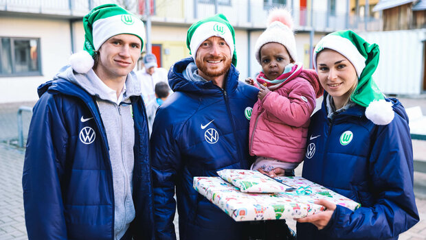 Die VfL Wolfsburg-Spieler Patrick Wimmer, Maximilian Arnold und Jule Brand machen ein Bild mit einem kleinen Kind und einem Geschenk.