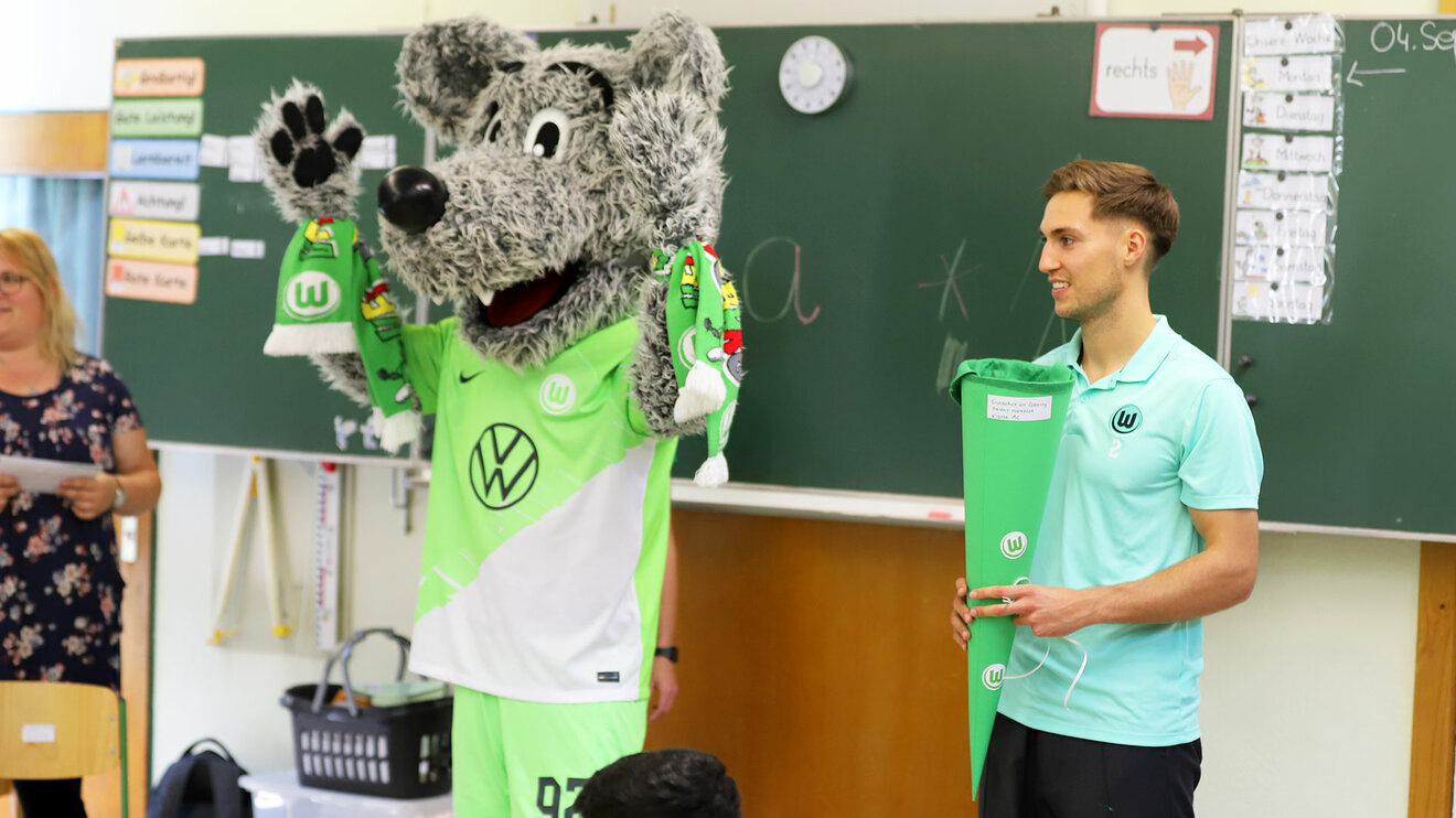 Wölfi und Kilian Fischer vom VfL Wolfsburg stehen in einem Klassenraum vor einer Tafel.