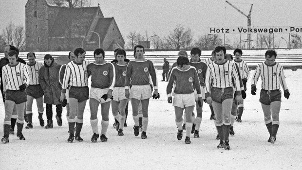 Ein historisches schwarz-weiß-Bild der Mannschaften des VfL Wolfsburg und des FC St. Pauli, die auf dem Spielfeld durch den Schnee laufen.