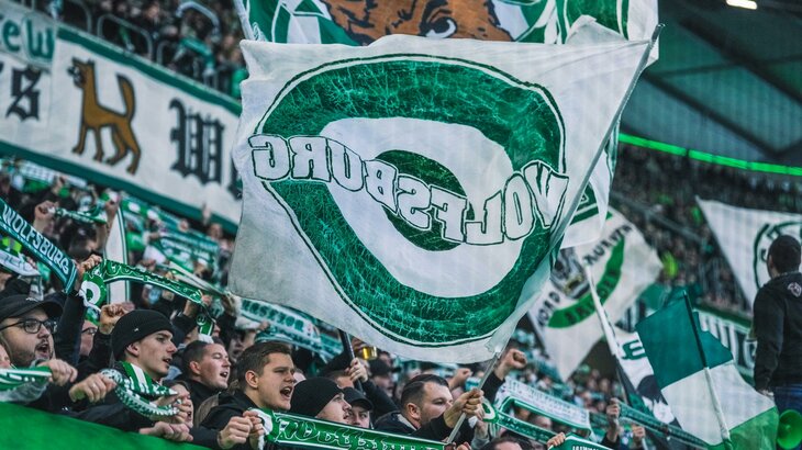 Ein Fan des VfL Wolfsburg wedelt in der Fankurve mit einer großen grün-weißen Fahne mit der Aufschrift Wolfsburg.