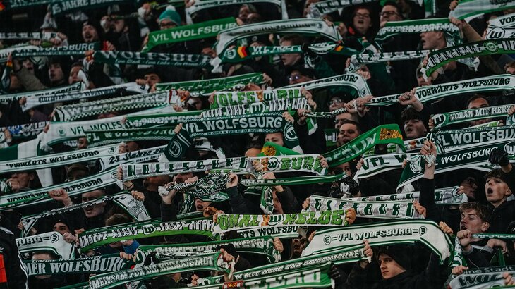 Fans des VfL-Wolfsburg halten ihren Schal auf der Tribüne in die Luft.