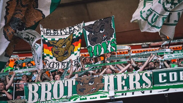 Es ist die Fanszene des VfL-Wolfsburg zu sehen. Zu erkennen ein Banner auf dem BROT und HERS steht und in der Mitte davon ein grimmiger Wolf. Außerdem werden Fahnen in die Luft geschwenkt.