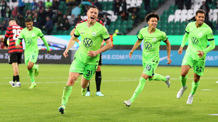 VfL Wolfsburg Spieler Yannik Gerhardt läuft jubelnd über das Spielfeld, im Hintergrund sieht man Paredes, der ebenfalls lächelt.