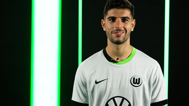 VfL Wolfsburg Spieler Konstantinos Koulierakis posiert im neuen Eventtrikot vor einer schwarzen Wand mit grünen Neon-Elementen.