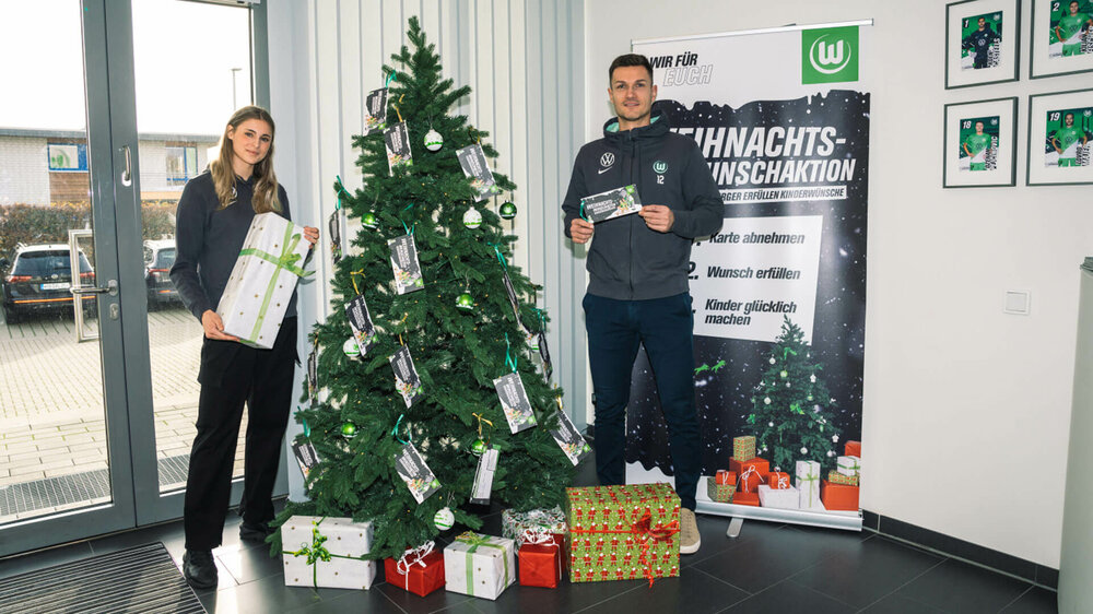 Die VfL-Wolfsburg-Spieler Jule Brand und Pavao Pervan stehen neben den Geschenken am Weihnachtsbaum.