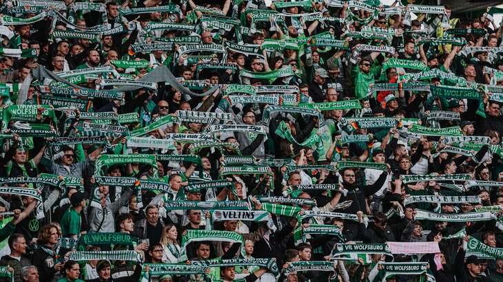 Fans des VfL-Wolfsburg stehen auf der Tribüne und halten ihre Schals in die Luft.