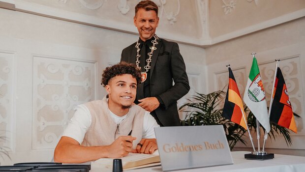 Der VfL-Wolfsburg-Spieler Kevin Paredes lacht in die Kamera. Vor ihm ein Aufsteller mit dem Schriftzug "goldenes Buch".
