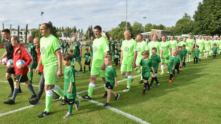 Die Teams laufen vor bunt gemischtem Publikum beim Jubiläumsspiel des VfL Wolfsburg mit den Einlaufkindern auf das Feld.