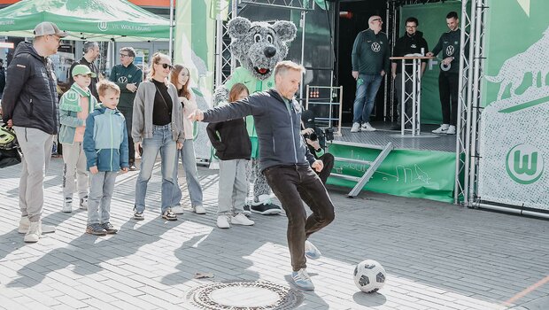 Ein Mann kickt mit dem linken Fuß den Ball Richtung Torwand. Hinter ihm stehen weitere Besucher und Wölfi und gucken zu. 