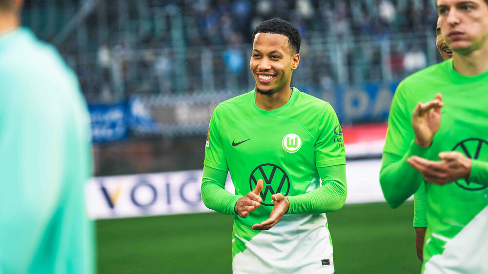 Der VfL-Wolfsburg-Spieler Aster Vranckx steht lachend auf dem Platz.