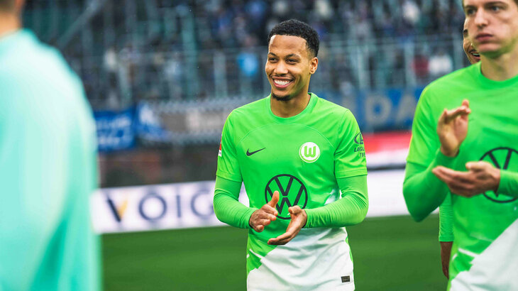 Der VfL-Wolfsburg-Spieler Aster Vranckx steht lachend auf dem Platz.