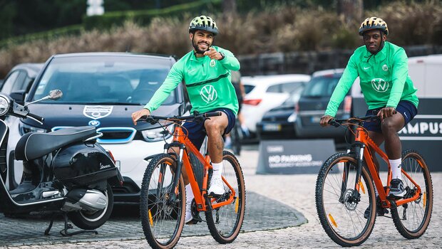 Die beiden VfL-Wolfsburg-Spieler Paulo Otavio und Jerome Roussillon fahren Fahrrad und lachen in die Kamera.