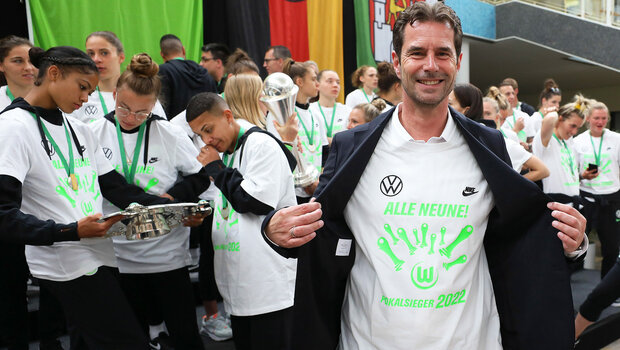 Sportlicher Leiter des VfL Wolfsburg Ralf Kellermann präsentiert sein T-Shirt mit der der Aufschrift "Alle Neune". Im Hintergrund sind die VfL-Frauen vor dem Rathaus zu sehen.