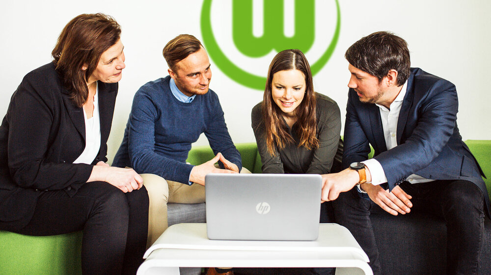 Mitarbeiter des VfL Wolfsburg diskutieren lächelnd vor einem Laptop Arbeitsinhalte. 
