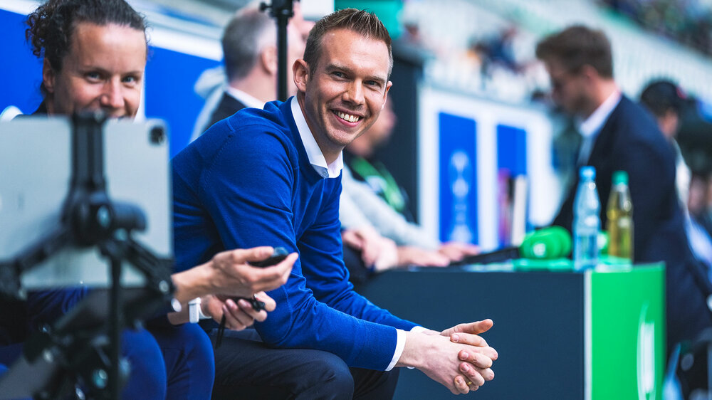 Tommy Stroot vom VfL Wolfsburg lehnt sich im Sitzen auf seinen Oberschenkeln ab und grinst.