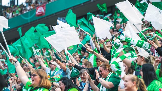 Die Fans des VfL Wolfsburg schwenken auf der Tribüne grün-weiße Fahnen.