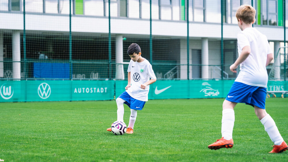 Zwei Jungen von der Fußballschule des VfL Wolfsburg spielen in blau-weißen Trikots Fußball.