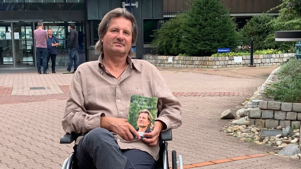 Philipp Hanf, Ansprechpartner der Krzysztof Nowak-Stiftung vom VfL Wolfsburg, sitzt im Rollstuhl und hält sein Buch in der Hand.