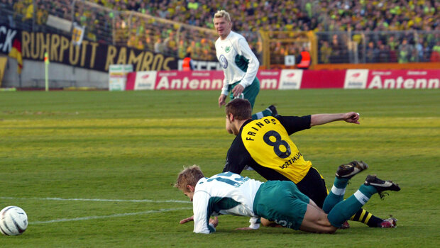 Ein VfL-Wolfsburg-Spieler liegt auf dem Boden, schräg über ihn geht ein BVB Spieler ebenfalls zu Boden. Vor ihnen ist der Spielball zu sehen. 