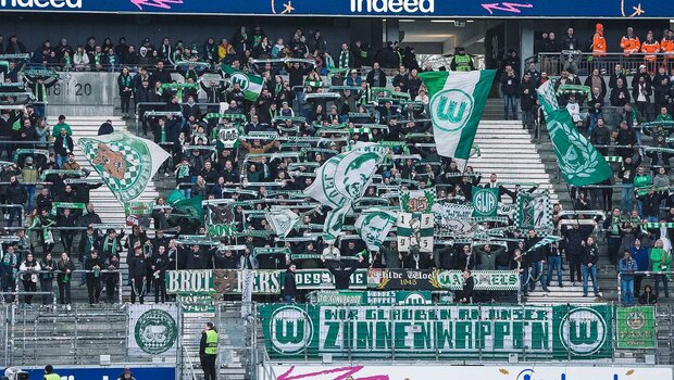 Die Fans des VfL Wolfsburg stehen auf der Tribüne nd halten Schals und Fahnen in die Höhe.