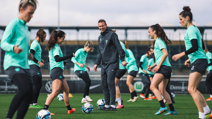 Tommy Stroot, Trainer der VfL Wolfsburg Frauen, steht bei einer Aufwärmübung mitten im Geschehen und lächelt seine Spielerin an.