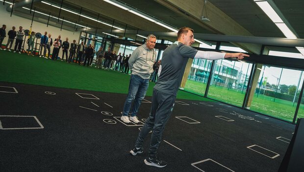 Ein Athletiktrainer zeigt einem VfL-Wolfsburg-Mitarbeiter im Kraftraum des neuen Funktionsgebäudes eine Bewegungsübung.
