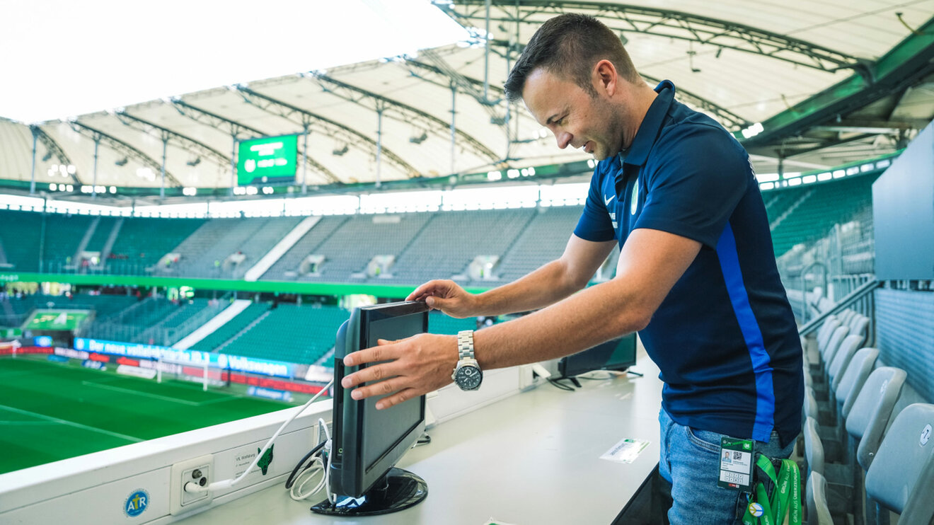 Ein Medienhelfer vom VfL Wolfsburg positioniert einen Monitor auf der Pressetribüne.
